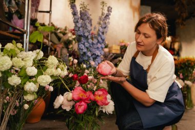 Önlüklü orta yaşlı kadın çiçekçi çiçekçide çiçek buketi için çiçek düzenliyor. Küçük bir yerel işletme. Dizinin bir parçası. Yumuşak odak