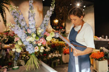 Önlüklü orta yaşlı kadın çiçekçi çiçek buketi için taze çiçek ayarlıyor çiçekçinin buketine bir kurdele bağlıyor. Küçük bir yerel işletme. Dizinin bir parçası. Yumuşak odak.