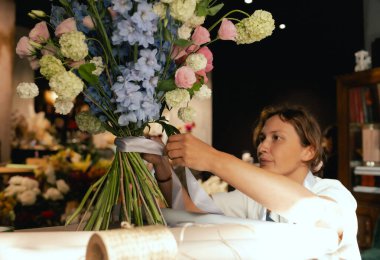 Önlüklü orta yaşlı kadın çiçekçi çiçek buketi için taze çiçek ayarlıyor çiçekçinin buketine bir kurdele bağlıyor. Küçük bir yerel işletme. Dizinin bir parçası. Yumuşak odak.