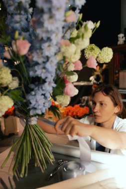 Önlüklü orta yaşlı kadın çiçekçi çiçek buketi için taze çiçek ayarlıyor çiçekçinin buketine bir kurdele bağlıyor. Küçük bir yerel işletme. Dizinin bir parçası. Yumuşak odak.