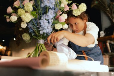 Önlüklü orta yaşlı kadın çiçekçi çiçek buketi için taze çiçek ayarlıyor çiçekçinin buketine bir kurdele bağlıyor. Küçük bir yerel işletme. Dizinin bir parçası. Yumuşak odak.