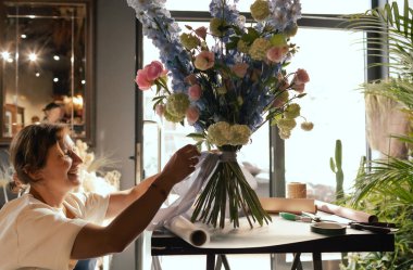 Önlüklü orta yaşlı kadın çiçekçi çiçek buketi için taze çiçek ayarlıyor çiçekçinin buketine bir kurdele bağlıyor. Küçük bir yerel işletme. Dizinin bir parçası. Yumuşak odak.