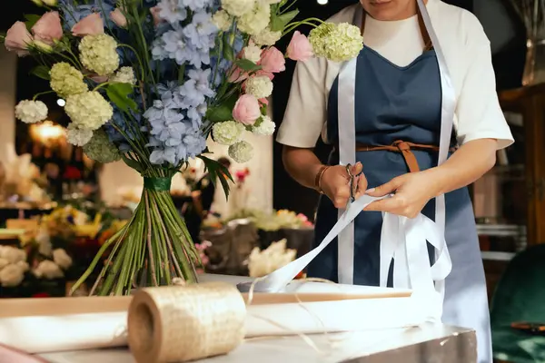 Önlüklü orta yaşlı kadın çiçekçi çiçek buketi için taze çiçek ayarlıyor çiçekçinin buketine bir kurdele bağlıyor. Küçük bir yerel işletme. Dizinin bir parçası. Yumuşak odak.