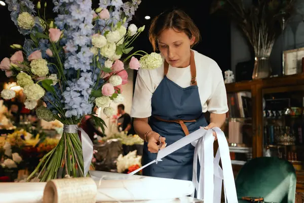 Önlüklü orta yaşlı kadın çiçekçi çiçek buketi için taze çiçek ayarlıyor çiçekçinin buketine bir kurdele bağlıyor. Küçük bir yerel işletme. Dizinin bir parçası. Yumuşak odak.