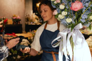 Çiçekçi: önlüklü kadın çiçekçi, çiçek buketi satıyor, müşteriler telefon aracılığıyla NFC terminali üzerinden ödeme yapıyor. Küçük bir yerel işletme. Cep telefonuna yakın çekim. Yumuşak odak