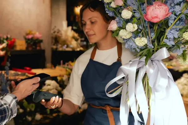 Çiçekçi: önlüklü kadın çiçekçi, çiçek buketi satıyor, müşteriler telefon aracılığıyla NFC terminali üzerinden ödeme yapıyor. Küçük bir yerel işletme. Cep telefonuna yakın çekim. Yumuşak odak