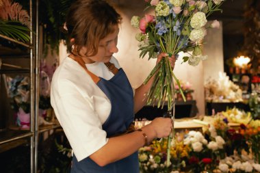 Önlüklü orta yaşlı kadın çiçekçi çiçekçide çiçek buketi için çiçek düzenliyor. Küçük bir yerel işletme. Dizinin bir parçası. Yumuşak odak