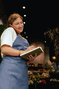 Önlüklü orta yaşlı kadın çiçekçi akıllı telefondan arıyor. Lojistiği ve teslimatı ayarlıyor, emirleri alıyor. Küçük bir yerel işletme. Dizinin bir parçası. Yumuşak odak