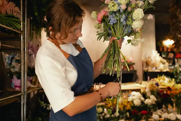 Önlüklü orta yaşlı kadın çiçekçi çiçekçide çiçek buketi için çiçek düzenliyor. Küçük bir yerel işletme. Dizinin bir parçası. Yumuşak odak