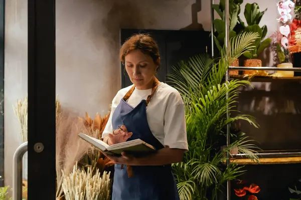 Önlüklü orta yaşlı kadın çiçekçi akıllı telefondan arıyor. Lojistiği ve teslimatı ayarlıyor, emirleri alıyor. Küçük bir yerel işletme. Dizinin bir parçası. Yumuşak odak