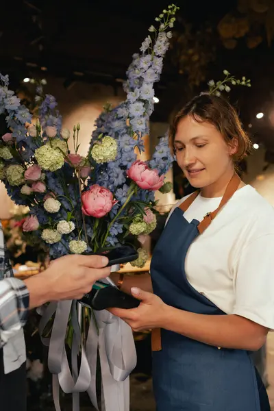 Çiçekçi: önlüklü kadın çiçekçi, çiçek buketi satıyor, müşteriler telefon aracılığıyla NFC terminali üzerinden ödeme yapıyor. Küçük bir yerel işletme. Cep telefonuna yakın çekim. Yumuşak odak