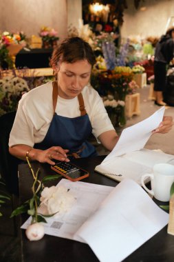 Orta yaşlı bir kadın, arka planda çeşitli bitkiler bulunan bir çiçekçide hesap makinesinin mali durumunu hesaplar. Lojistiği ve teslimatı ayarlıyor, emirleri alıyor. Küçük yerel işletme