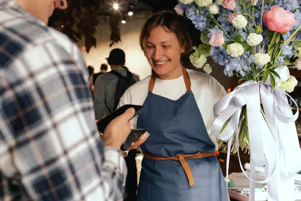 Çiçekçi: önlüklü kadın çiçekçi, çiçek buketi satıyor, müşteriler telefon aracılığıyla NFC terminali üzerinden ödeme yapıyor. Küçük bir yerel işletme. Cep telefonuna yakın çekim. Yumuşak odak
