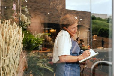 Önlüklü orta yaşlı kadın çiçekçi akıllı telefondan arıyor. Lojistiği ve teslimatı ayarlıyor, emirleri alıyor. Küçük bir yerel işletme. Dizinin bir parçası. Yumuşak odak