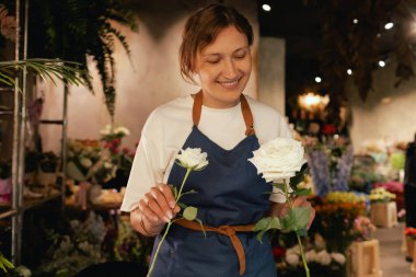Önlüklü orta yaşlı kadın çiçekçi çiçekçide çiçek buketi için çiçek düzenliyor. Küçük bir yerel işletme. Dizinin bir parçası. Yumuşak odak