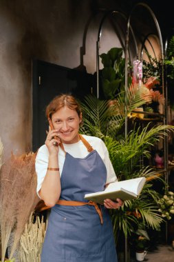 Önlüklü orta yaşlı kadın çiçekçi akıllı telefondan arıyor. Lojistiği ve teslimatı ayarlıyor, emirleri alıyor. Küçük bir yerel işletme. Dizinin bir parçası. Yumuşak odak