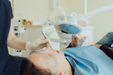 Genç bir dişçiye dişçi muayenesinde gülümseyen olgun bir kadın. Klinikte randevu. Diş beyazlatma, temizlik ya da oral, hastanede ağız ve sakız bakımı. Yumuşak odaklanma. Dizinin bir parçası. Yakın plan..