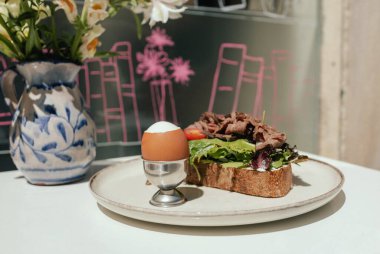 Etli sandviç, yumurta ve taze yeşillik. Güneşli açık kafede, ahşap masada sunulan lezzetli mahsul kahvaltısı. Bir serinin parçası.