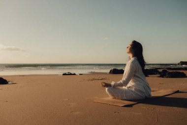 Beyaz elbiseli Asyalı kadın meditasyon için yoga yapıyor ve deniz kenarında sağlıklı olmak için dinleniyor. Sağlık hizmeti egzersizi ve yoga konsepti. Solo turizm. Bir serinin parçası.