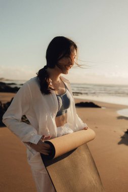 Okyanus kıyısında yoga minderli beyaz elbiseli Asyalı mutlu kadın. Sağlık egzersizleri ve yoga kavramı. Sadece turizm. Dizinin bir parçası.