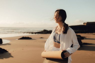 Okyanus kıyısında yoga minderli beyaz elbiseli Asyalı mutlu kadın. Sağlık egzersizleri ve yoga kavramı. Sadece turizm. Dizinin bir parçası.
