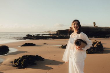 Okyanus kıyısında yoga minderli beyaz elbiseli Asyalı mutlu kadın. Sağlık egzersizleri ve yoga kavramı. Sadece turizm. Dizinin bir parçası.