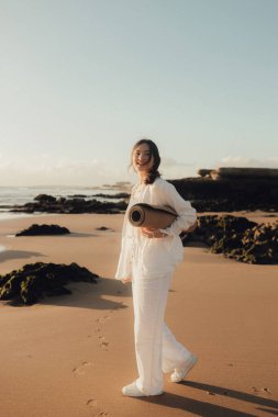 Okyanus kıyısında yoga minderli beyaz elbiseli Asyalı mutlu kadın. Sağlık egzersizleri ve yoga kavramı. Sadece turizm. Dizinin bir parçası.
