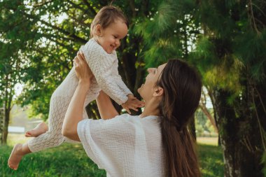 Güneşli bir parkta bebeğiyle çimlerin üzerinde oynayan mutlu genç bir anne. Aile bağları, erken öğrenme, ebeveynlik, aşk, açık hava yaşam tarzı, çocukluk gelişimi, otantik gerçek yaşam anı. Bir serinin parçası.