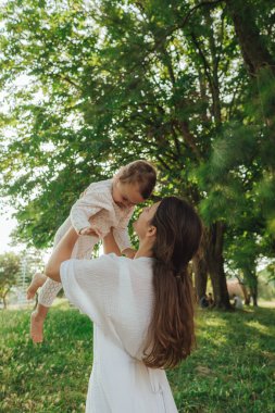 Güneşli bir parkta bebeğiyle çimlerin üzerinde oynayan mutlu genç bir anne. Aile bağları, erken öğrenme, ebeveynlik, aşk, açık hava yaşam tarzı, çocukluk gelişimi, otantik gerçek yaşam anı. Bir serinin parçası.