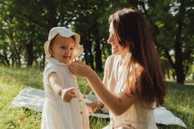 Güneşli bir parkta bebeğiyle çimlerin üzerinde oynayan mutlu genç bir anne. Aile bağları, erken öğrenme, ebeveynlik, aşk, açık hava yaşam tarzı, çocukluk gelişimi, otantik gerçek yaşam anı. Bir serinin parçası.