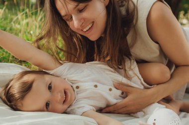 Güneşli bir parkta bebeğiyle çimlerin üzerinde oynayan mutlu genç bir anne. Aile bağları, erken öğrenme, ebeveynlik, aşk, açık hava yaşam tarzı, çocukluk gelişimi, otantik gerçek yaşam anı. Bir serinin parçası.