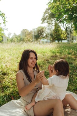 Güneşli bir parkta bebeğiyle çimlerin üzerinde oynayan mutlu genç bir anne. Aile bağları, erken öğrenme, ebeveynlik, aşk, açık hava yaşam tarzı, çocukluk gelişimi, otantik gerçek yaşam anı. Bir serinin parçası.