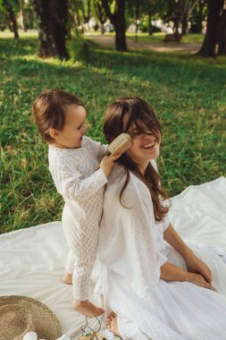 Güneşli bir parkta bebeğiyle çimlerin üzerinde oynayan mutlu genç bir anne. Aile bağları, erken öğrenme, ebeveynlik, aşk, açık hava yaşam tarzı, çocukluk gelişimi, otantik gerçek yaşam anı. Bir serinin parçası.
