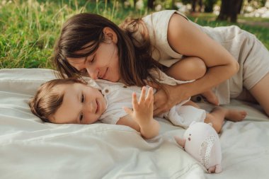 Güneşli bir parkta bebeğiyle çimlerin üzerinde oynayan mutlu genç bir anne. Aile bağları, erken öğrenme, ebeveynlik, açık hava yaşam tarzı, çocukluk gelişimi, otantik gerçek yaşam anı. Bir serinin parçası