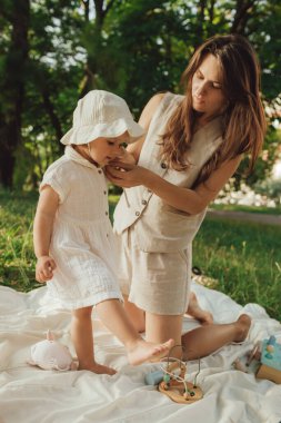 Güneşli bir parkta bebeğiyle çimlerin üzerinde oynayan mutlu genç bir anne. Aile bağları, erken öğrenme, ebeveynlik, açık hava yaşam tarzı, çocukluk gelişimi, otantik gerçek yaşam anı. Bir serinin parçası