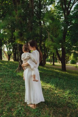 Güneşli bir parkta bebeğiyle çimlerin üzerinde oynayan mutlu genç bir anne. Aile bağları, erken öğrenme, ebeveynlik, açık hava yaşam tarzı, çocukluk gelişimi, otantik gerçek yaşam anı. Bir serinin parçası