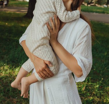 Güneşli bir parkta bebeğiyle çimlerin üzerinde oynayan mutlu genç bir anne. Aile bağları, erken öğrenme, ebeveynlik, açık hava yaşam tarzı, çocukluk gelişimi, otantik gerçek yaşam anı. Bir serinin parçası