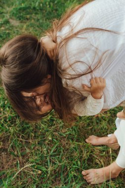 Güneşli bir parkta bebeğiyle çimlerin üzerinde oynayan mutlu genç bir anne. Aile bağları, erken öğrenme, ebeveynlik, açık hava yaşam tarzı, çocukluk gelişimi, otantik gerçek yaşam anı. Bir serinin parçası
