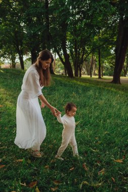 Güneşli bir parkta bebeğiyle çimlerin üzerinde oynayan mutlu genç bir anne. Aile bağları, erken öğrenme, ebeveynlik, açık hava yaşam tarzı, çocukluk gelişimi, otantik gerçek yaşam anı. Bir serinin parçası