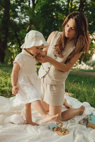 Güneşli bir parkta bebeğiyle çimlerin üzerinde oynayan mutlu genç bir anne. Aile bağları, erken öğrenme, ebeveynlik, açık hava yaşam tarzı, çocukluk gelişimi, otantik gerçek yaşam anı. Bir serinin parçası