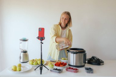 Orta yaşlı bir kadın ev yapımı meyve derisi hazırlıyor. Mutfağında taze meyve dilimleyip sağlıklı bir şeyler atıştırıyor ve telefonundan yayın yapıyor. Dizinin bir parçası. Kullanıcı tarafından oluşturulan içerik.