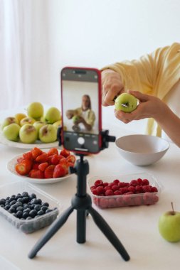 Orta yaşlı bir kadın ev yapımı meyve derisi hazırlıyor. Mutfağında taze meyve dilimleyip sağlıklı bir şeyler atıştırıyor ve telefonundan yayın yapıyor. Dizinin bir parçası. Kullanıcı tarafından oluşturulan içerik.