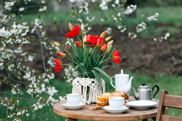 田舎風の庭とお茶会 静物画 カップ ケーキ 赤いチューリップの花瓶 ロイヤリティフリーのストック画像