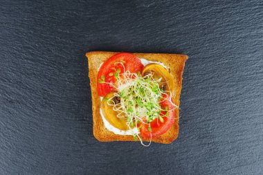 Yeşil yonca filizleri, taze ve kurutulmuş domatesler kızarmış kepekli ekmeğin üzerinde siyah kayalık arka planda.