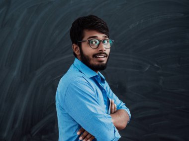Mavi gömlekli genç bir Hintli öğrenci okul tahtasının önünde kollarını kavuşturmuş poz veriyor..