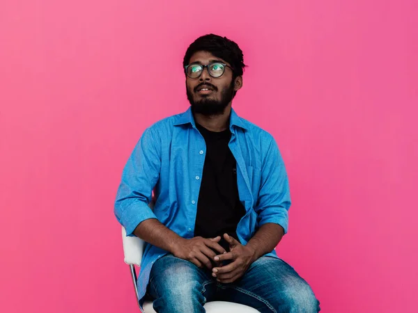 一位身穿蓝色衬衫 戴着眼镜的印度青年学生坐在粉色背景的前面 — 图库照片