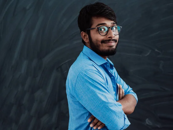 一名身穿蓝色衬衫 双手举着眼镜的印度青年学生在学校黑板前交叉站立 — 图库照片