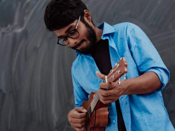 身穿蓝色衬衫 戴着眼镜 在学校黑板前弹奏吉他的印度年轻人 — 图库照片