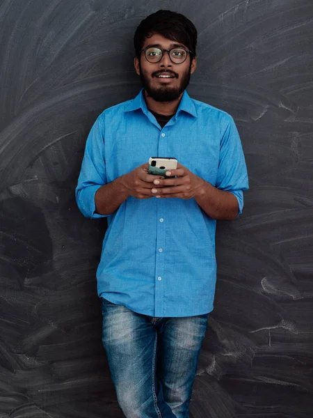 インド人の笑顔の青いシャツと眼鏡を使用してスマートフォンや学校の黒板の背景にポーズ — ストック写真