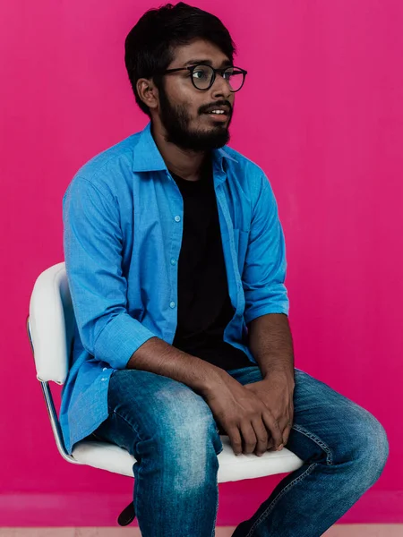 一位身穿蓝色衬衫 戴着眼镜的印度青年学生坐在粉色背景的前面 — 图库照片